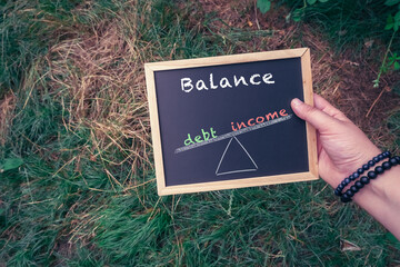 Hand holding chalkboard with balance concept illustrating debt versus income on grass background