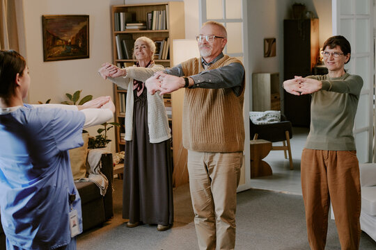 Senior Caucasian man and two senior Caucasian women standing with arms extended forward, following instructions from young adult Caucasian woman in medical uniform during exercise session