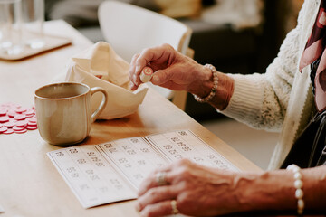 Senior Caucasian woman sitting at table playing bingo, holding numbered bingo chip in hand, marking...