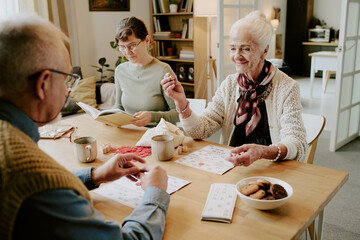 Senior Caucasian woman smiling while playing bingo with senior Caucasian man at table, middle aged Caucasian woman reading book in background, group engaging in leisure activity indoors
