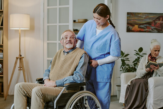 Fototapeta Caucasian senior man with disability sitting in wheelchair being assisted by young adult Hispanic woman caregiver while elderly Caucasian woman reading book in background