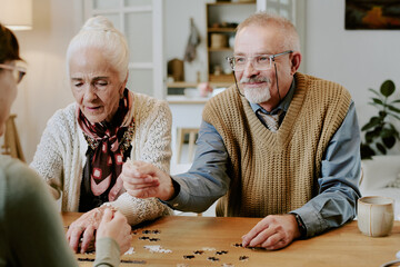 Senior Caucasian woman and senior Caucasian man sitting at table assembling puzzle pieces together with another , both focusing on activity and interacting with puzzle