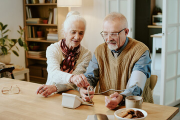 Senior Caucasian woman assisting senior Caucasian man measuring blood pressure with digital monitor at table, both focusing on device, bookshelf and open door visible in background