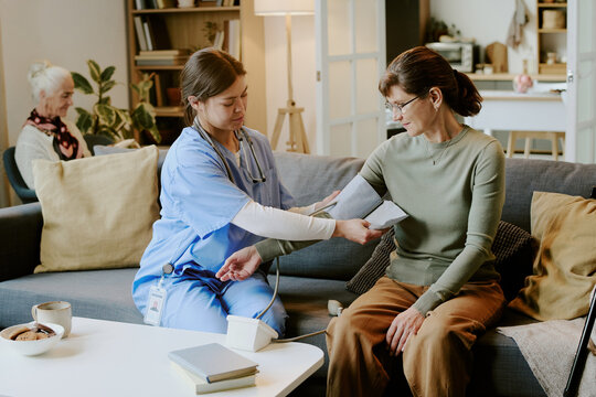 Young adult Hispanic female nurse measuring blood pressure of middle aged Caucasian woman sitting on sofa, while senior Caucasian woman sitting in background reading book in living room - Powered by Adobe