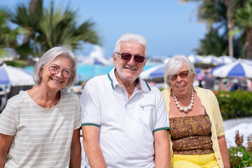 Three cheerful senior friends enjoy a sunny day at a seaside resort in a vibrant beach atmosphere with umbrellas, palm trees, and blue sky, expressing relaxation, friendship, and vacation happiness