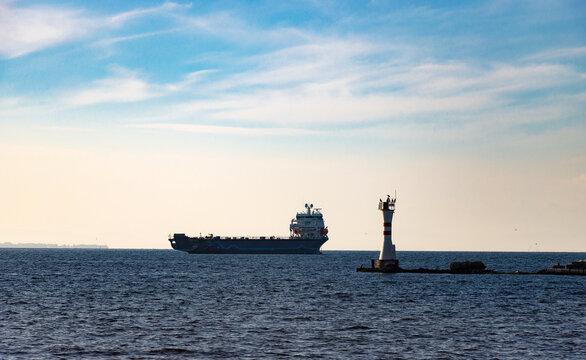Cargo ship passing near red-striped lighthouse on rocky outcrop—captured in maritime composition with calm sea, perched birds, and clear daylight atmosphere. Cloudy Daylight, Transportation Concept.