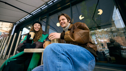 Friends laughing and drinking hot coffee on cafe bench in winter weather. Concept of lifestyle...