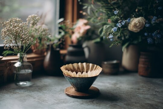 Coffee pour over dripper with paper filter rests on wooden coaster, flowers blurred in background. - Powered by Adobe