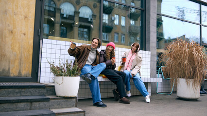 Smiling friends sitting with takeaway drinks on tiled outdoor bench. Concept of cafe ads, urban winter life, social bloggers, wellbeing stories, financial care themes and mobile ordering.