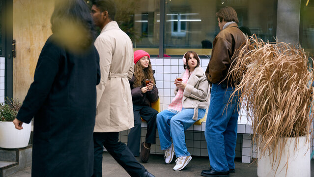 Friends chatting on outdoor bench near winter cafe exterior. Concept of coffee shop promos, winter travel, social lifestyle, event banners, wellbeing warmth, and mobile delivery ads.