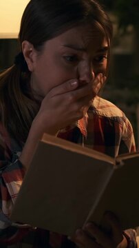 Anxious young woman reading scary moment in a book, then sighing with relief and smiling. Emotional female reader reacting to plot twist at home at night. Vertical video.