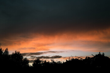 Obraz premium Dramatic orange and dark sky at sunset or sunrise above a tree line.