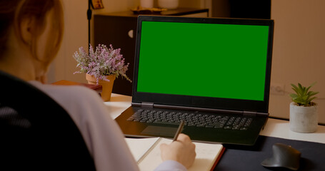 A woman focuses on her laptop with a green screen, jotting down notes in her notebook in a cozy home office environment. She is engaged in her tasks effectively.
