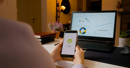 Businesswoman confirming online payment on smartphone with laptop showing graphs in background