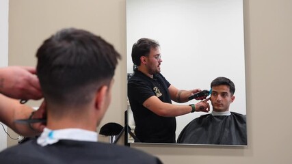 Lively hairdresser using a trimmer to cut customer’s hair while chatting in a modern barbershop mirror reflection