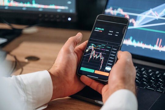 Against the laptop. Detailed close up view of man's hands that is holding smartphone with stock market crypto information - Powered by Adobe