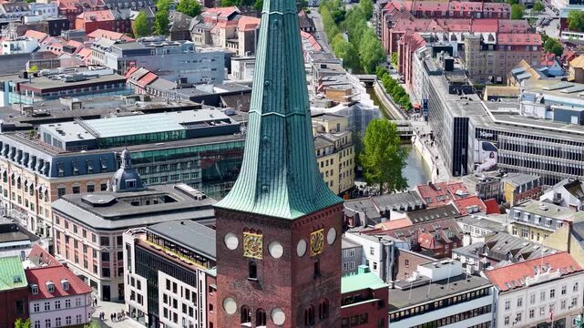 Aerial close‑up of Aarhus city center highlighting a historic church tower with a green spire, clock faces and surrounding colorful rooftops.
