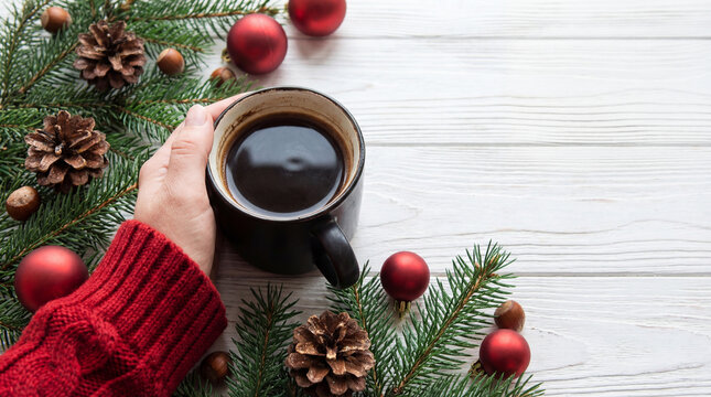 Hand holding warm coffee mug on festive christmas table with evergreen branches, pine cones, red ornaments and cozy sweater sleeve - Powered by Adobe