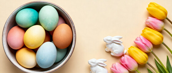 Obraz premium colorful pastel easter eggs in a ceramic bowl with yellow and pink tulips and white bunny figurines on a light background
