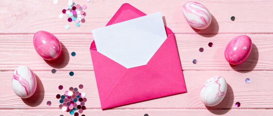 Pink envelope with blank card surrounded by decorative marbled easter eggs and confetti on pastel wooden background
