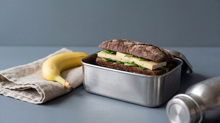 Healthy sandwich with fresh cheese and greens in metal lunchbox, banana and reusable bottle on neutral background for on the go meals
