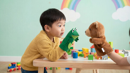 Curious young boy playing with frog and bear hand puppets at a wooden table in a colorful playroom with rainbow wall art