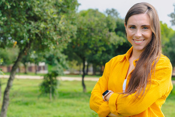 Confident woman wearing yellow shirt smiling outdoors in green park with crossed arms and copy...