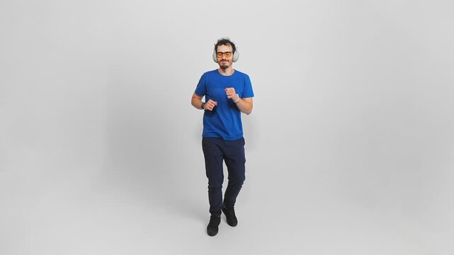 Cheerful young adult guy is walking straight looking at camera and listening to music in earphones. Caucasian guy in casual clothes is enjoying his favorite song and dancing moving on white background