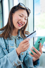 Happy chinese woman on tram holding a credit card and smartphone. Wearing sunglasses and denim...