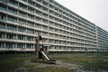 Old, abandoned playground with a rusty slide against a long, brutalist apartment building. A grim, post-soviet urban landscape on an overcast day.