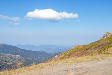Mountain landscape with dirt road, dry grass, autumn foliage, rocky outcrop and white cloud under blue sky