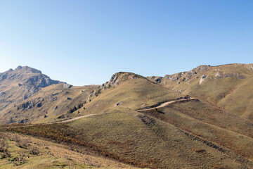 Mountain landscape with dirt road, grassy hills, rocky outcrops and sparse vegetation under clear blue sky