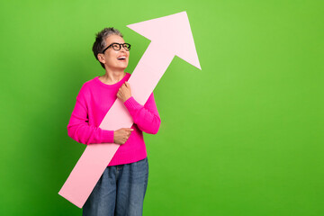 Mature woman in pink sweater smiles while holding a large pink arrow against a green background symbolizing growth progress and a positive lifestyle for advertising