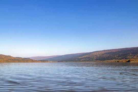 Mountain lake with gentle ripples, autumn forested hills and distant ridges under clear blue sky - Powered by Adobe
