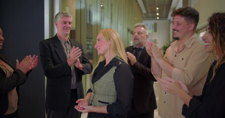 Smiling woman receiving applause from diverse coworkers in office hallway, group of professionals...