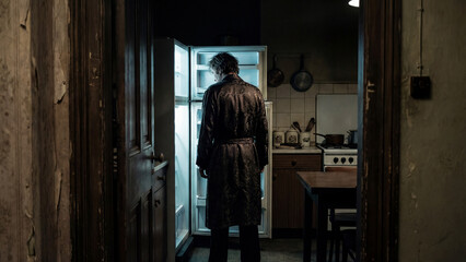 A lonely man in a coat stands in front of an open refrigerator in a dark, old kitchen. A moody, atmospheric scene of poverty, loneliness, and despair.