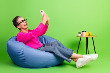 A spirited mature lady in a pink sweater relaxes on a blue bean bag while taking a selfie with her smartphone in a bright green studio setting