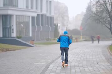 Jogger running along fog-covered paved urban path on cold morning as mist softens background...