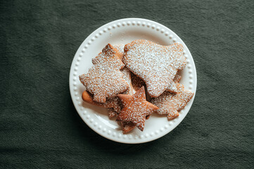 A white plate with Christmas gingerbread cookies sprinkled with powdered sugar on a dark green textured tablecloth.