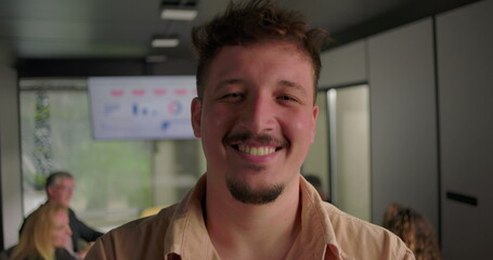 Smiling young man looking at camera in office, friendly expression with coworkers engaged at laptops in modern workspace