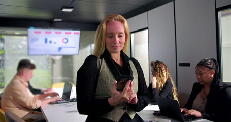 Serious businesswoman looks at smartphone with concern during office meeting, focused team working on laptops, data visualization screen in background