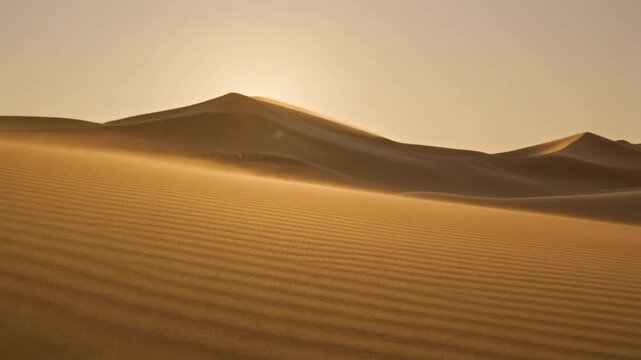 Golden sand dunes ripple under a hazy sky as wind blows sand from the crest at sunset.