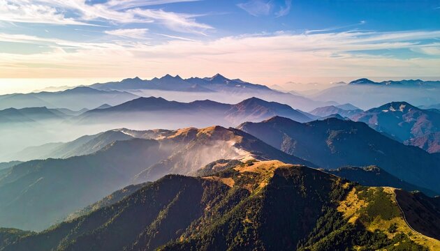 Aerial view of a mountain range with layers of peaks, fog, and a blue sky. The scene is bathed in soft, natural light.