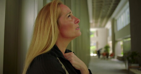 Fototapeta premium Side view of woman lowering her gaze with hand near chest in corporate corridor with modern seating and soft natural light through windows