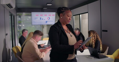 Smiling African American businesswoman holding digital tablet in office, confident leadership presence as team works at conference table in background