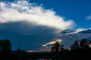Dramatic stormy sky with bright white clouds contrasting dark blue heavy rain clouds near sunset silhouettes of trees reflecting on calm water surface