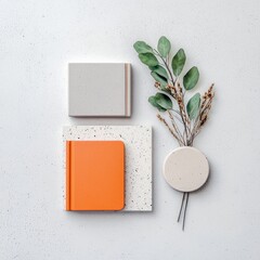 A top-down shot showcases an orange notebook, a gray square, a textured square, and a small vase with greenery, all arranged on a white textured surface.