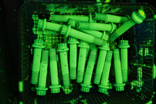 Fluorescent Penetrant Testing of Industrial Threaded Bolts Under UV Light. Green Glowing Fasteners in Wire Basket Showing NDT Quality Control Inspection for Defect Detection Process