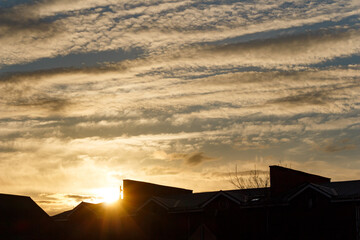 Brilliant golden sunset flares behind dark building silhouettes with striking layered clouds streaking across the sky