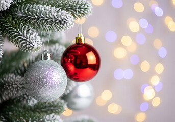 Close-up of a frosted Christmas tree featuring a glossy red bauble and a silver ornament. Bright holiday background with shimmering bokeh lights.